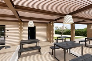 Een terras buiten met tafels en banken, een bakstenen muur en uitzicht op groen.