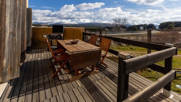 Una terraza de madera con una mesa, sillas, una parrilla y vistas a montañas nevadas.