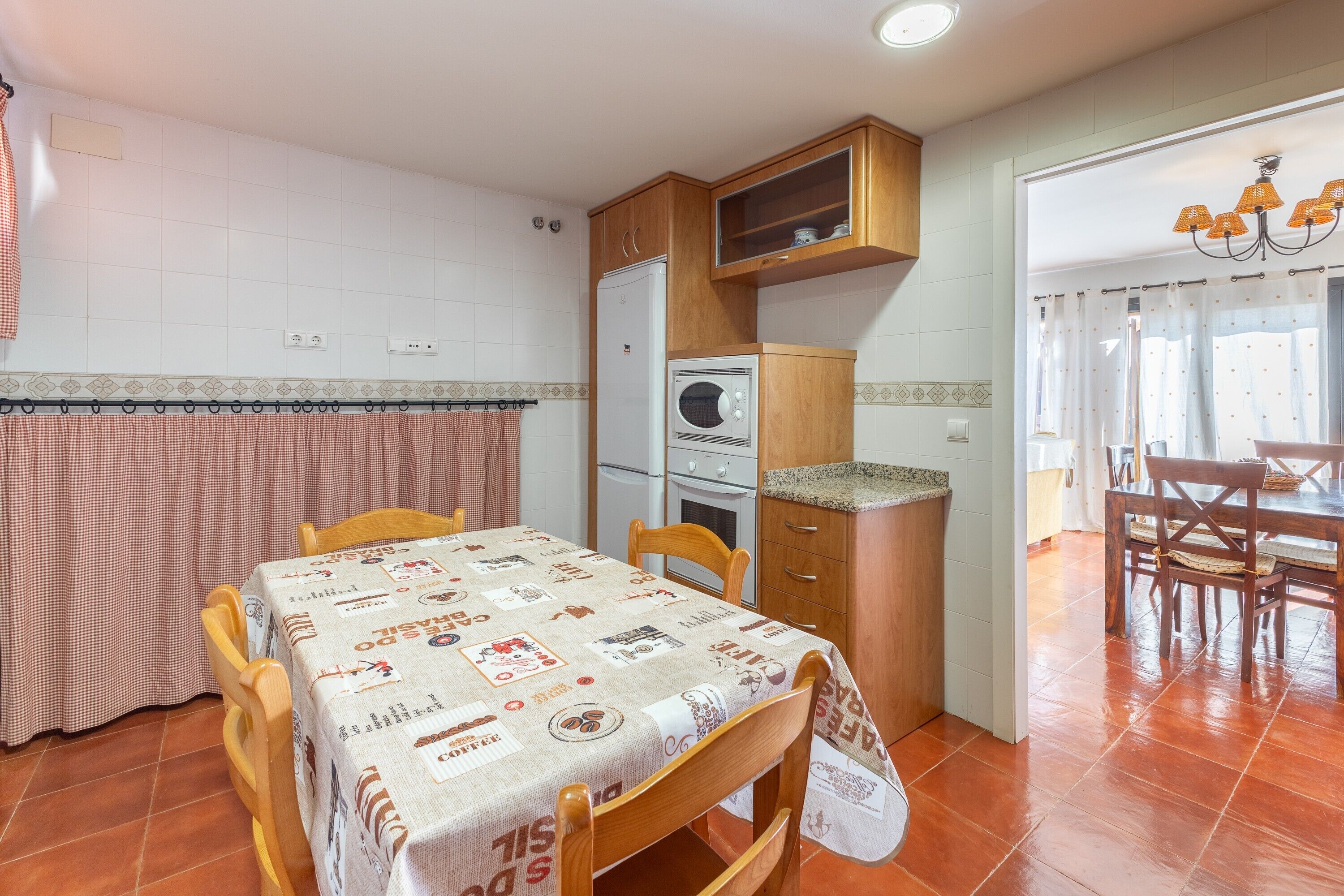 Une cuisine compacte avec un coin repas, comprenant une table recouverte d’une nappe, des chaises en bois, un réfrigérateur, un four et un four à micro-ondes.