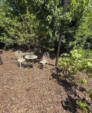 Un jardin avec deux chaises en métal et une table, entouré d’un feuillage dense et d’un poteau en bois.