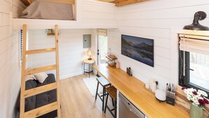 A compact, modern kitchen with a wooden countertop, bar stools, and a dining area. A loft bed with a blanket is visible, and a large window provides natural light.