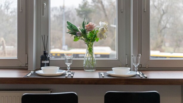 Una mesa de comedor dispuesta con platos blancos, una copa de vidrio transparente y un jarrón con flores.