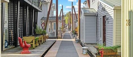 A narrow wooden pathway flanked by residential buildings with a metal bridge overhead.