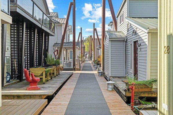 A narrow wooden pathway flanked by residential buildings with a metal bridge overhead.