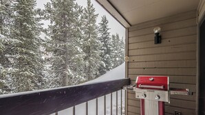 Een balkon met een barbecue, houten wanden en besneeuwde bomen op de achtergrond.