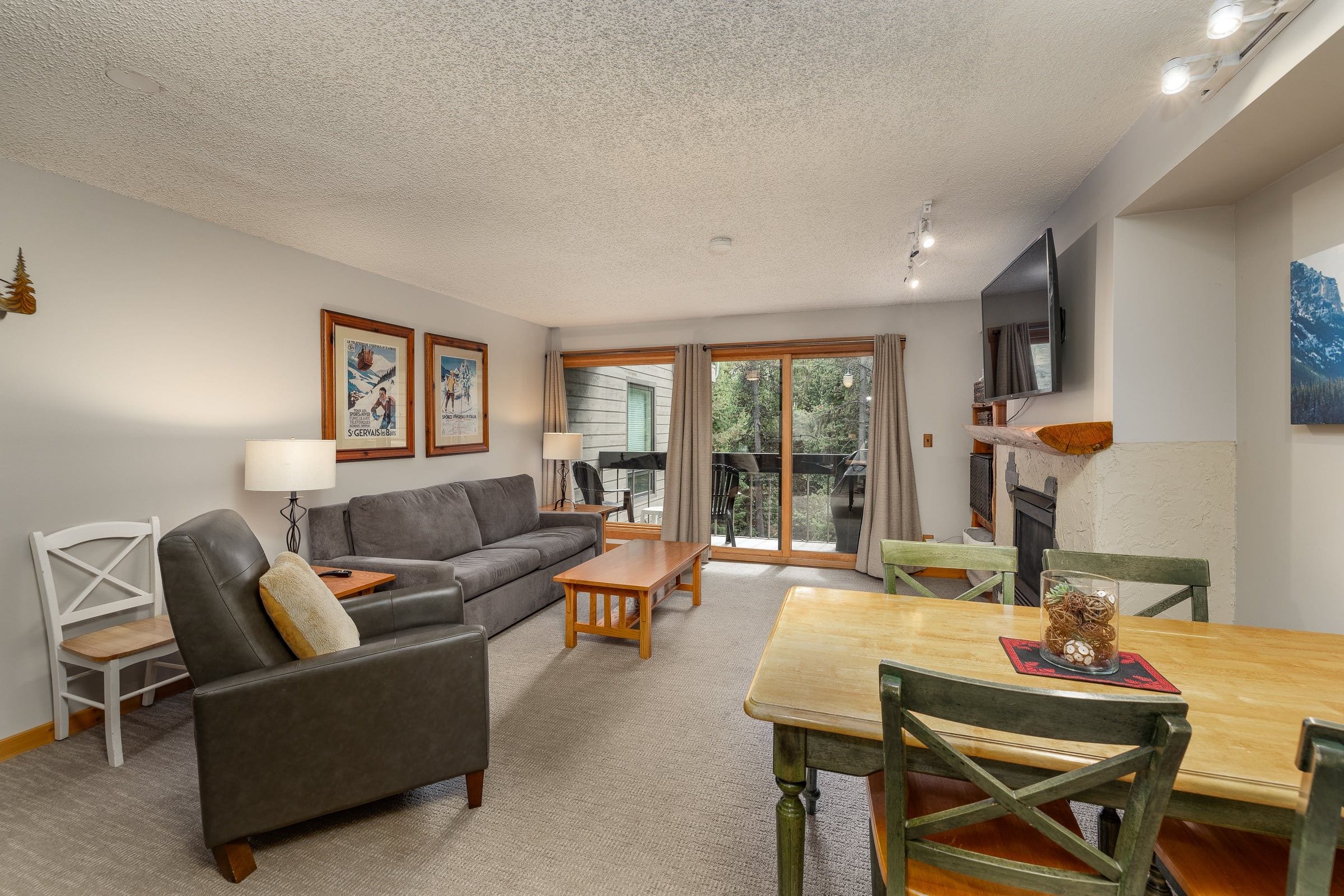 A living room with a grey sofa, a wooden coffee table, and a dining table with chairs. There are two framed pictures on the wall, a television mounted on the wall, and a fireplace.