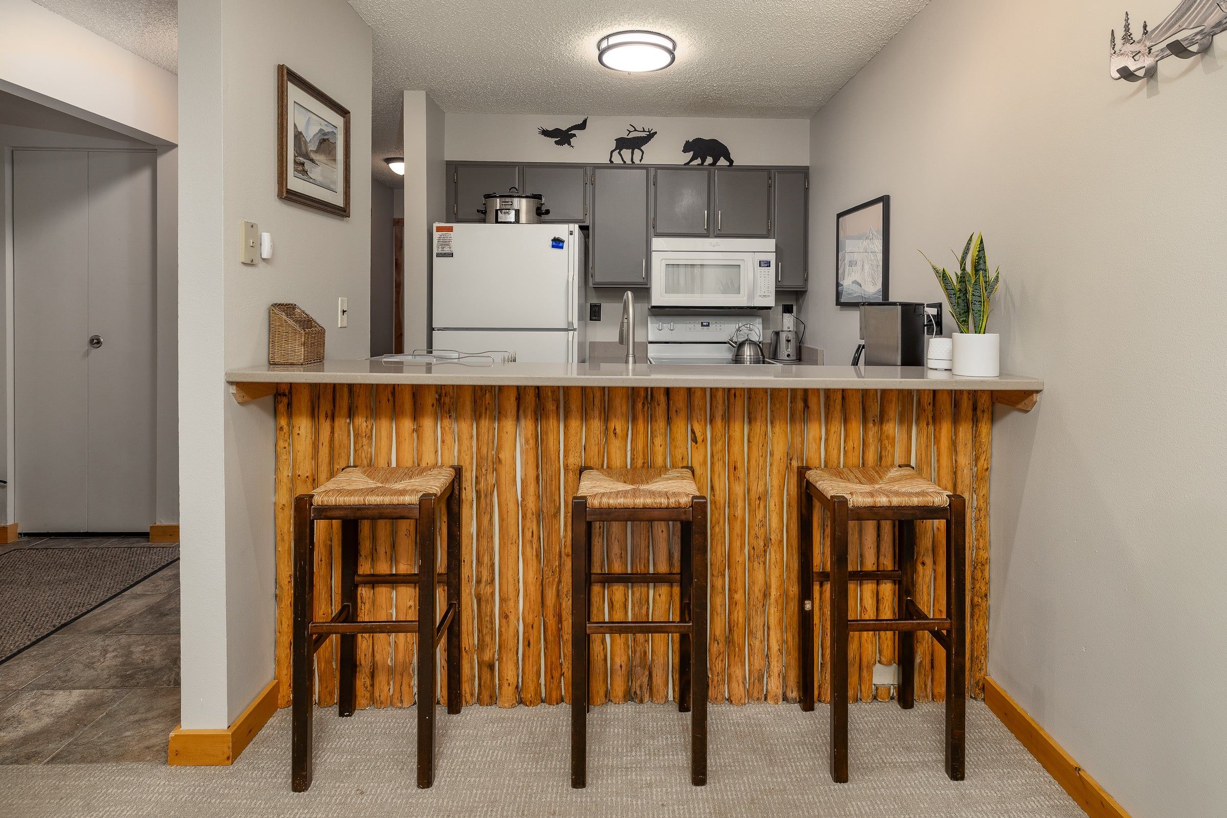 A kitchen with a wooden bar area, three bar stools, a microwave, a refrigerator, and a sink.