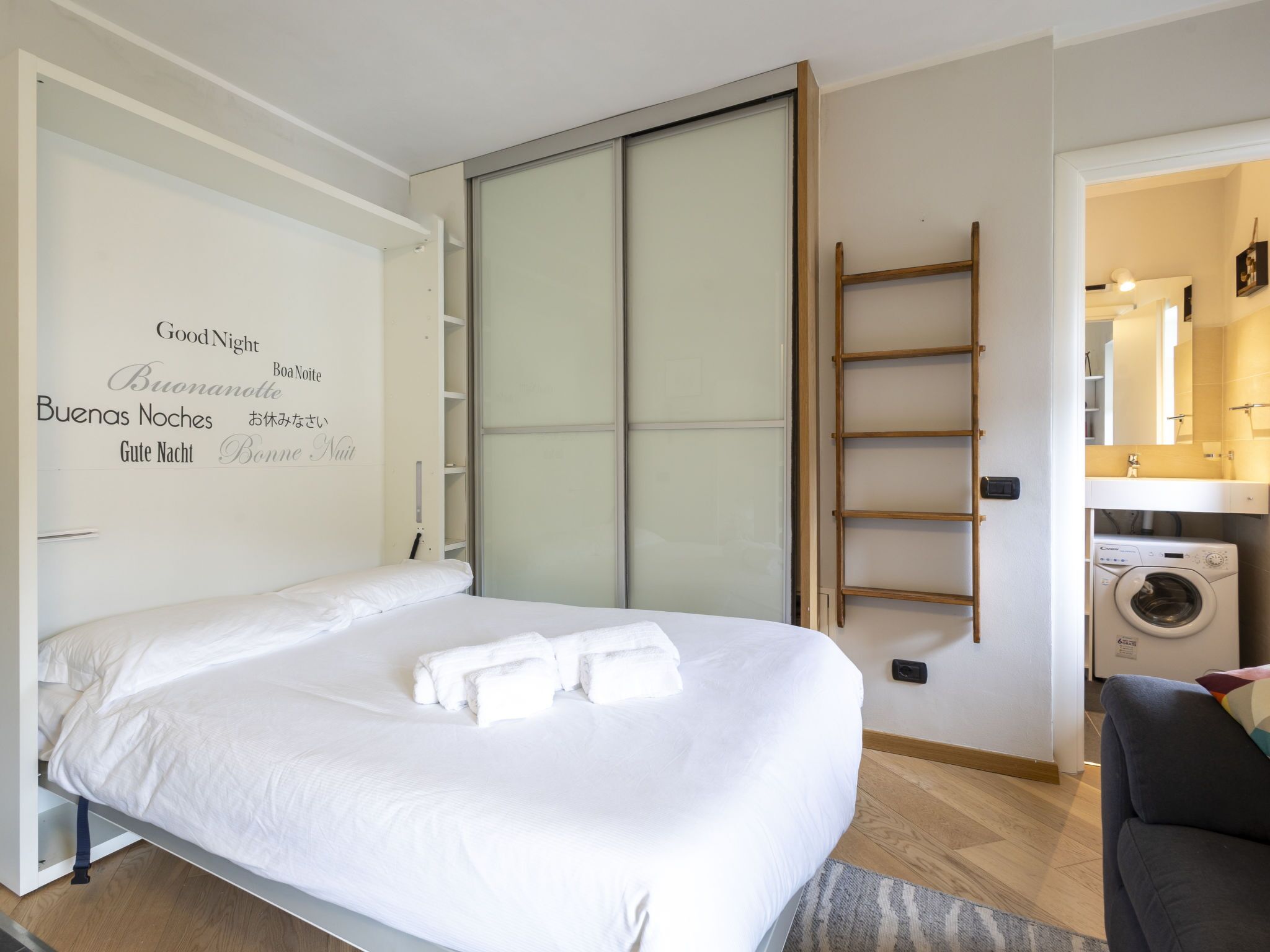 Ein Schlafzimmer mit Bett, Schrank, Waschmaschine und einem durchgehenden Türrahmen einsehbarer Badezimmer.