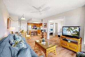 A living room with a sofa, coffee table, and television. There is a kitchen in the background with wooden cabinets and a dining area with chairs.