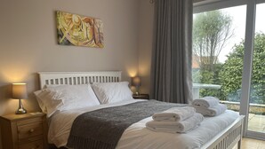 A bedroom with a bed, bedside tables, a lamp, a painting, and a view of greenery outside.