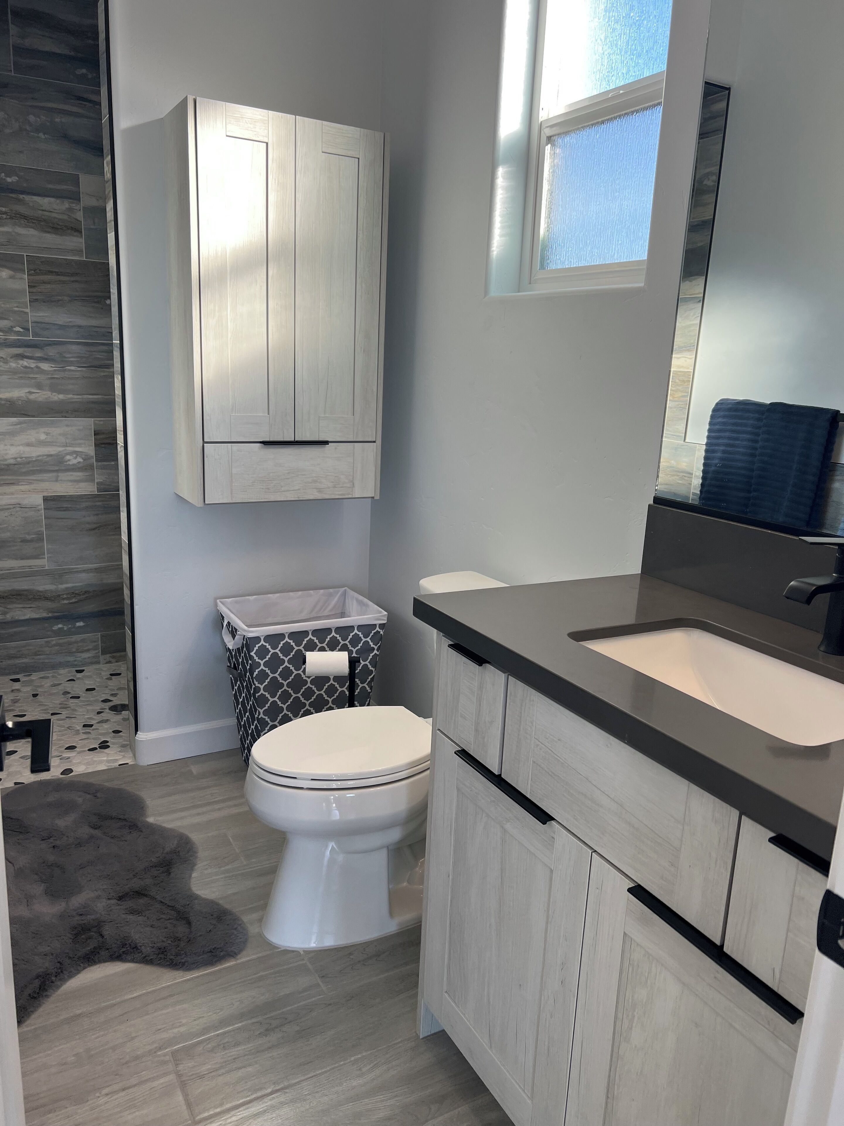 A modern bathroom with a grey and white color scheme, featuring a toilet, a sink with a black countertop, and a wooden cabinet above the sink.