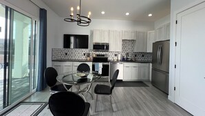 A modern kitchen with a glass dining table, black chairs, a stainless steel refrigerator, and a flat-screen TV.