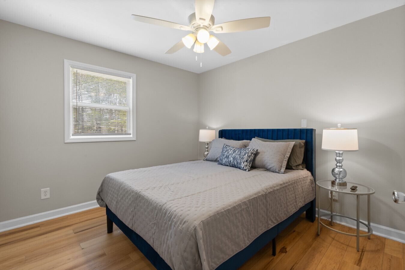 A bedroom with a bed, a ceiling fan, a window with blinds, and a bedside table with a lamp.