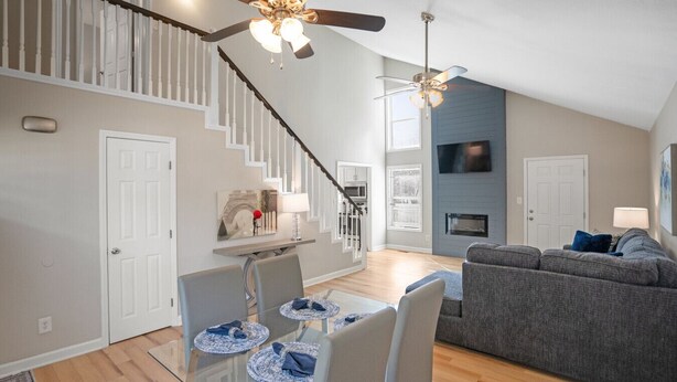 A modern living room with a gray sofa, a glass dining table with blue placemats, a staircase with white railings, and a fireplace. - Tranquil Retreat Near Lake Norman – Cozy, Spacious & Perfect for Relaxation! (Denver)