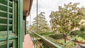 Un balcón con barandilla de metal que da a un jardín con diversas plantas y árboles.