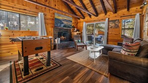 Interior acogedor de una cabaña de troncos, con una mesa de fútbol de mesa, chimenea, sofá y ventanas grandes.