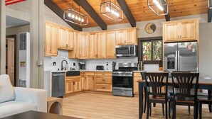 A modern kitchen with wooden cabinets, stainless steel appliances, and a dining area with black chairs. - Timber Wolf Tavern - Walk to Slopes, Game Room, Pool Table (Big Bear Lake)