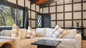 A modern living room with a sectional sofa, a wooden coffee table, and a large window with a view of trees. - Timber Wolf Tavern - Walk to Slopes, Game Room, Pool Table (Big Bear Lake)