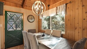 A wooden interior with a dining table, chairs, a chandelier, a green door, and a window with curtains.