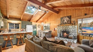 A cozy cabin interior with a stone fireplace, wooden ceiling beams, a dining area, and a bar.