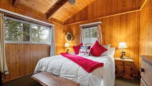 A wooden cabin bedroom with a large bed, two bedside lamps, a wooden bench, and a window with curtains.