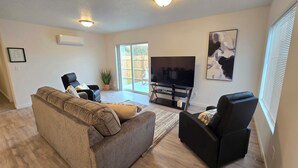 A living room with a large sofa, a black armchair, a flat-screen TV on a stand, a potted plant, and a sliding glass door leading to an outdoor area. - Hill Base Modern (Umatilla)