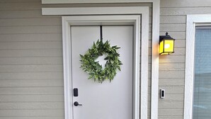 A front door with a wreath, a 'hello' mat, and a wall-mounted light fixture. - Hill Base Modern (Umatilla)