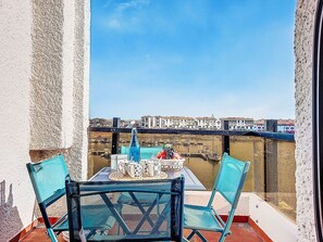 Un balcon avec une table dressée pour deux personnes, des chaises et une vue sur un port bordé de bâtiments.