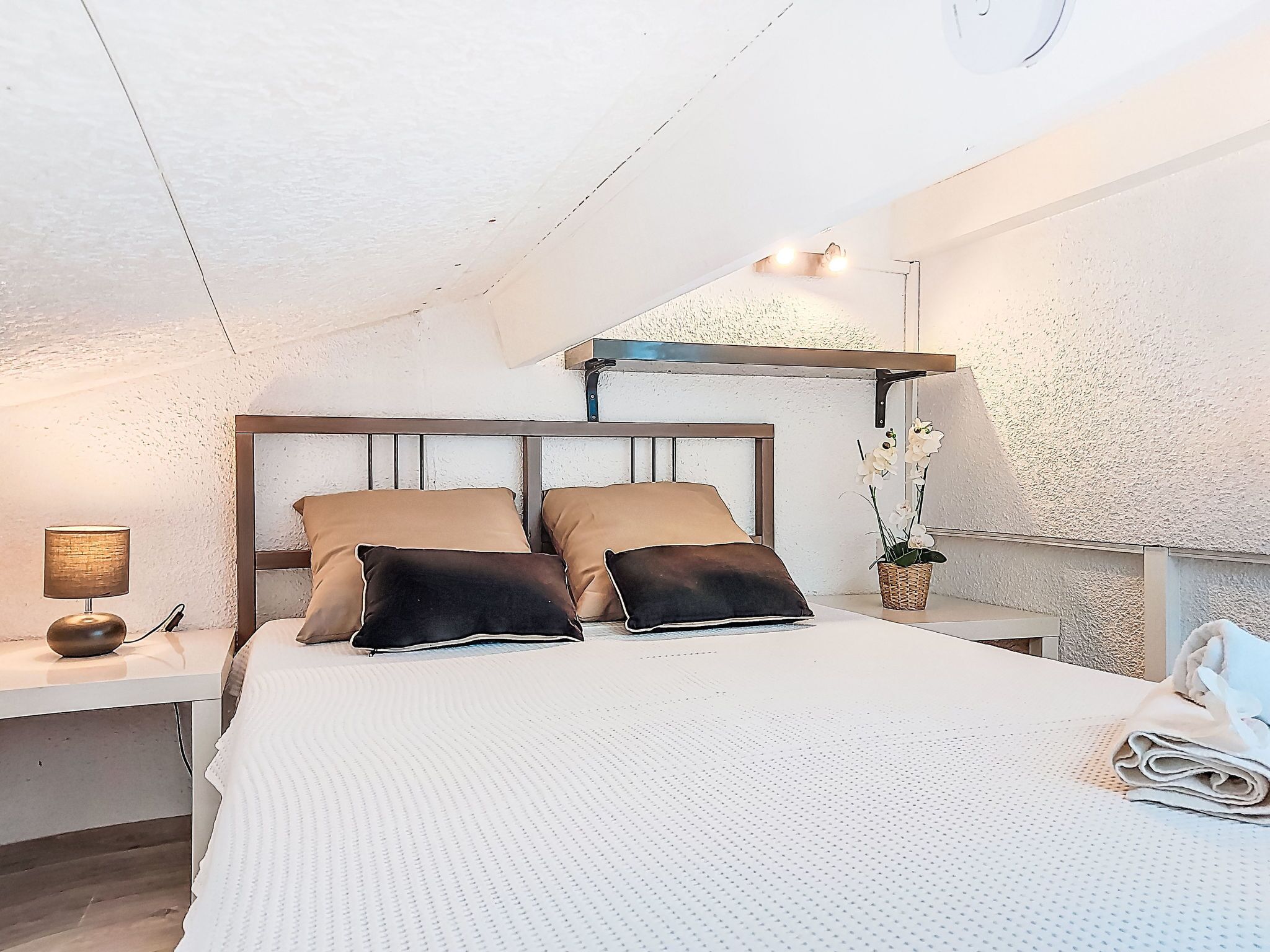 Un dormitorio con una cama, almohadas, una lámpara de noche, una mesita, un estante en la pared y una planta en maceta.
