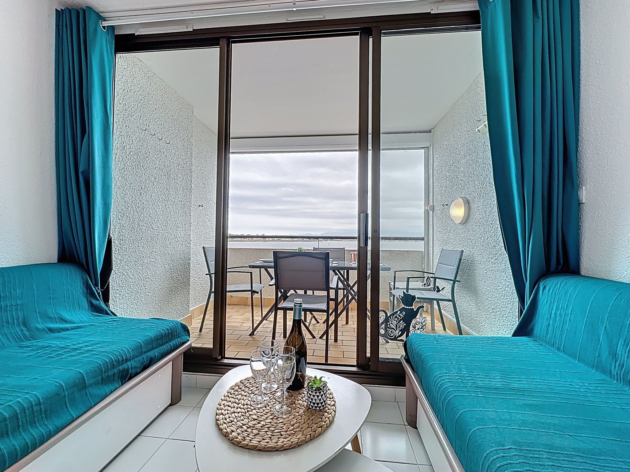 A balcony with a view of the sea, furnished with two teal sofas, a table with wine glasses, and a chair.
