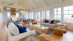 A spacious living room with a high ceiling, large windows, and a modern kitchen in the background. - SBT115 Private Pool, Pickleball Court, Sauna, Boardwalk to the Beach (Port Aransas)