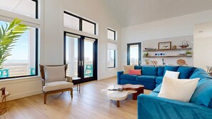 A spacious living room with a blue sofa, a wooden chair, and a coffee table. Large windows offer a view of the beach. - Shared Resort Style Pools, Boardwalk to the Beach, Beautiful Views (Port Aransas)
