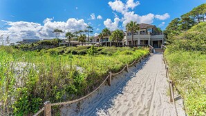 Una propiedad frente a la playa con un camino de arena que conduce a un edificio de varios pisos, rodeado de vegetación y palmeras.