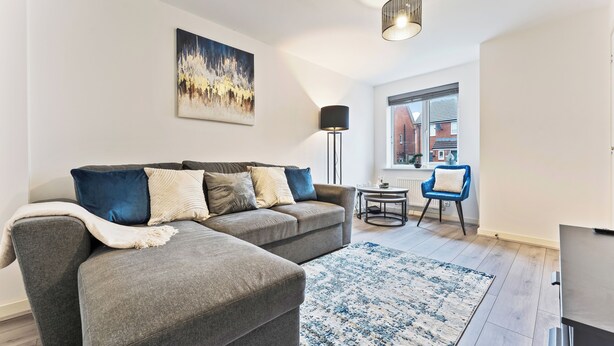 A modern living room with a grey sectional sofa, a blue chair, a small table, and a large window with a view of buildings. - City Centre House.Parking.Sofa Bed.WIFI (Coventry)