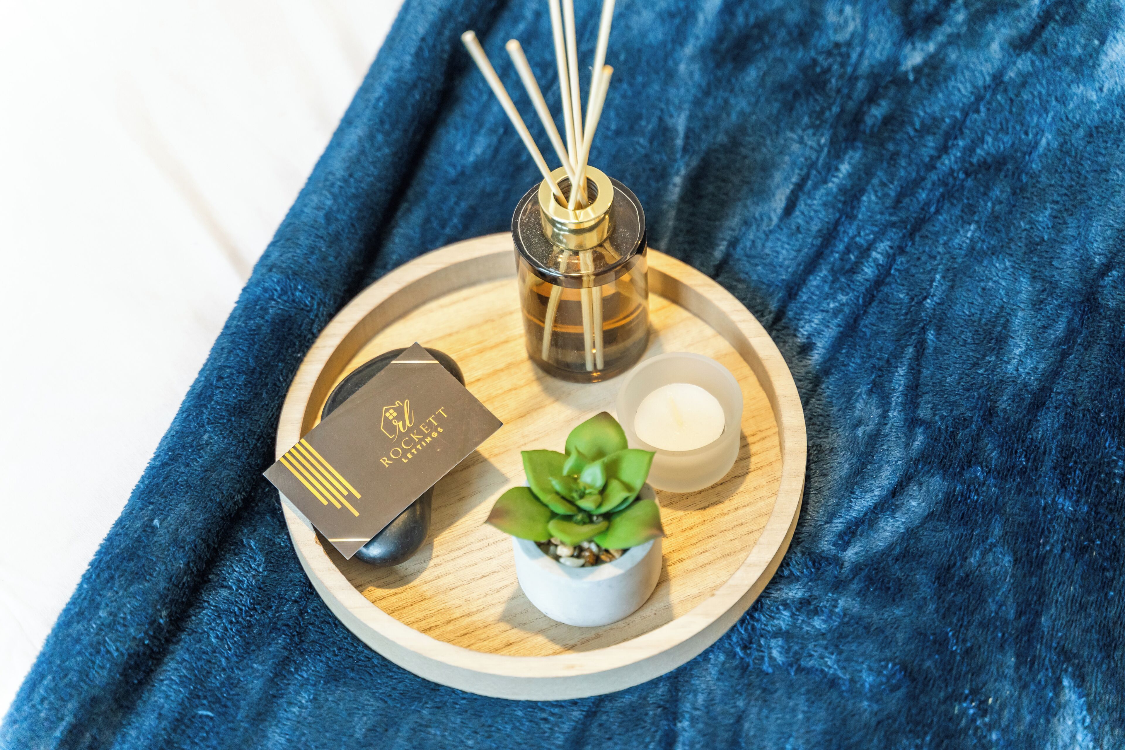 A tray with a small potted plant, a candle, reed diffusers, and a gift card on a blue fabric background.