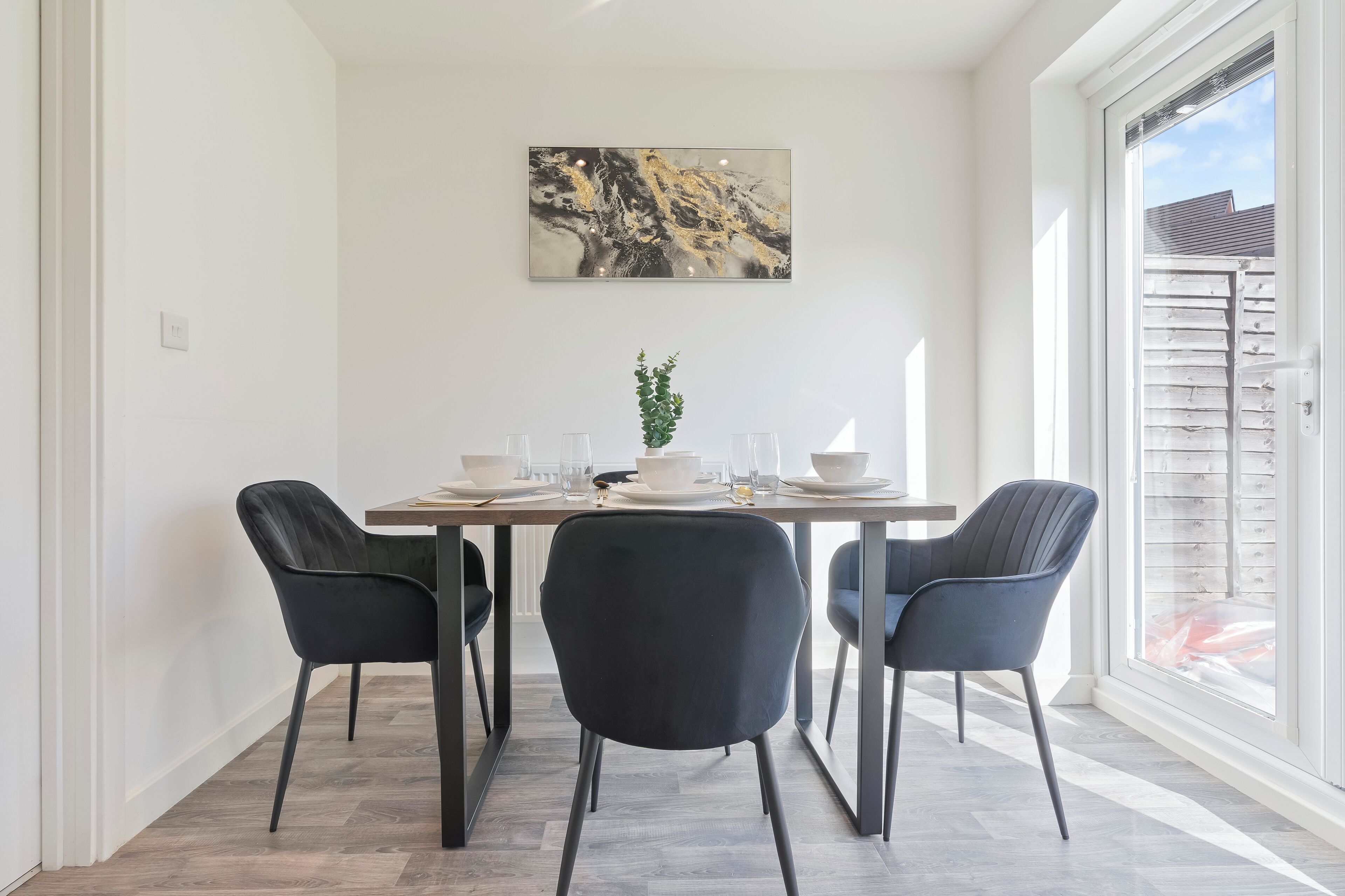 A dining area with a table set for four, two black chairs, a plant, and a painting on the wall.