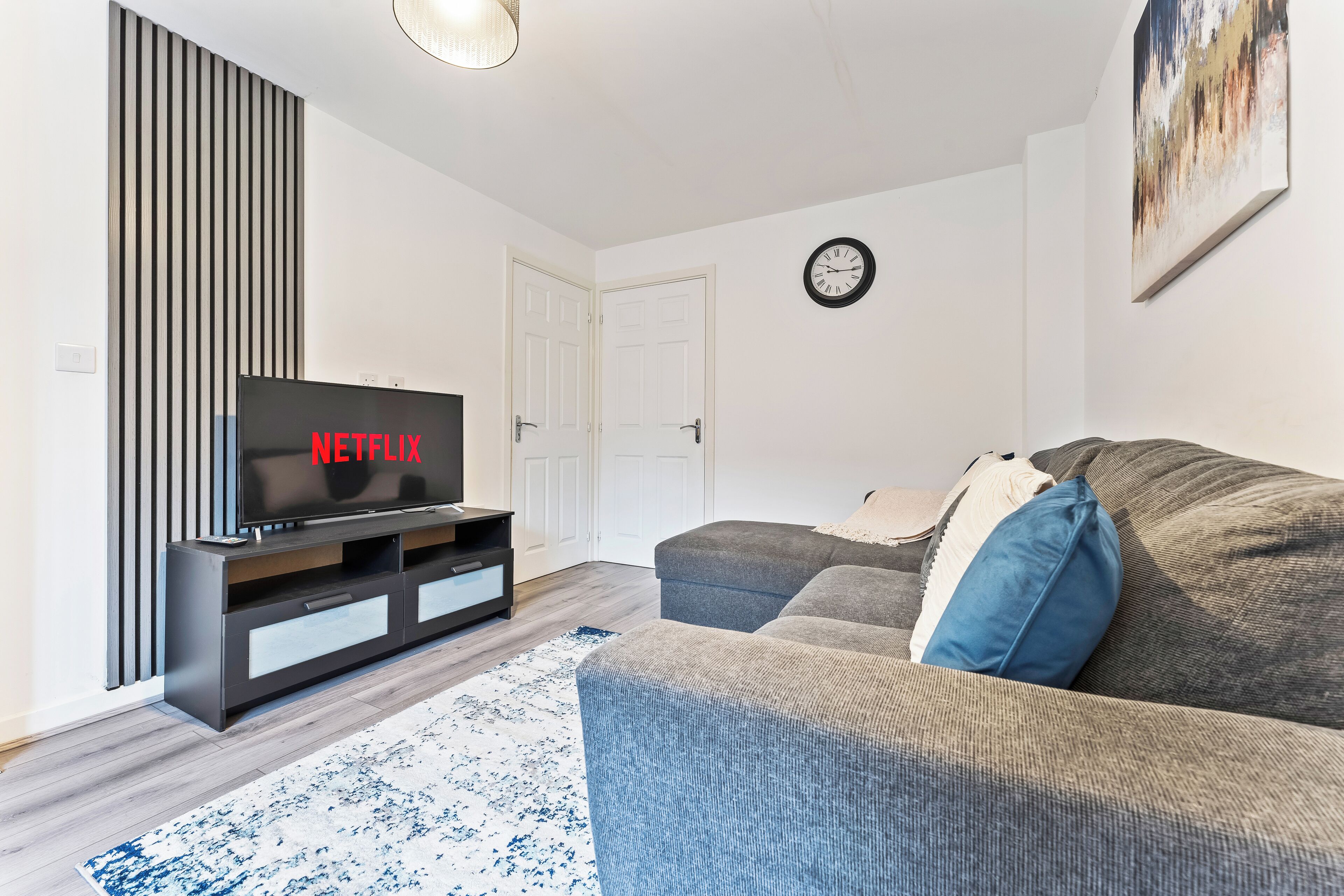 A modern living room with a flat-screen TV displaying 'NETFLIX', a grey sofa, a patterned rug, and a wall clock.