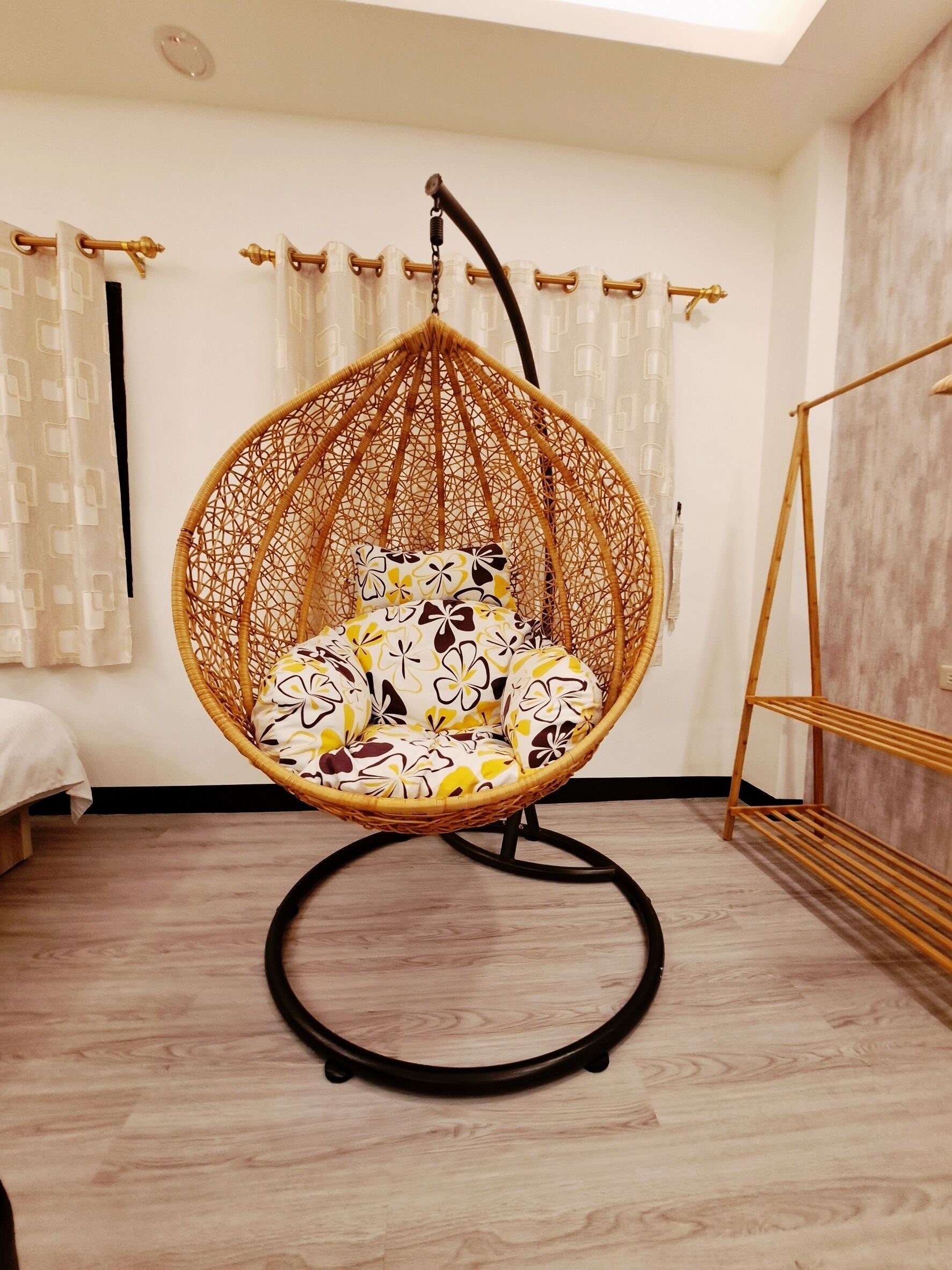 A room with a hanging chair with a floral cushion, a wooden floor, and a bed with a white bedspread.