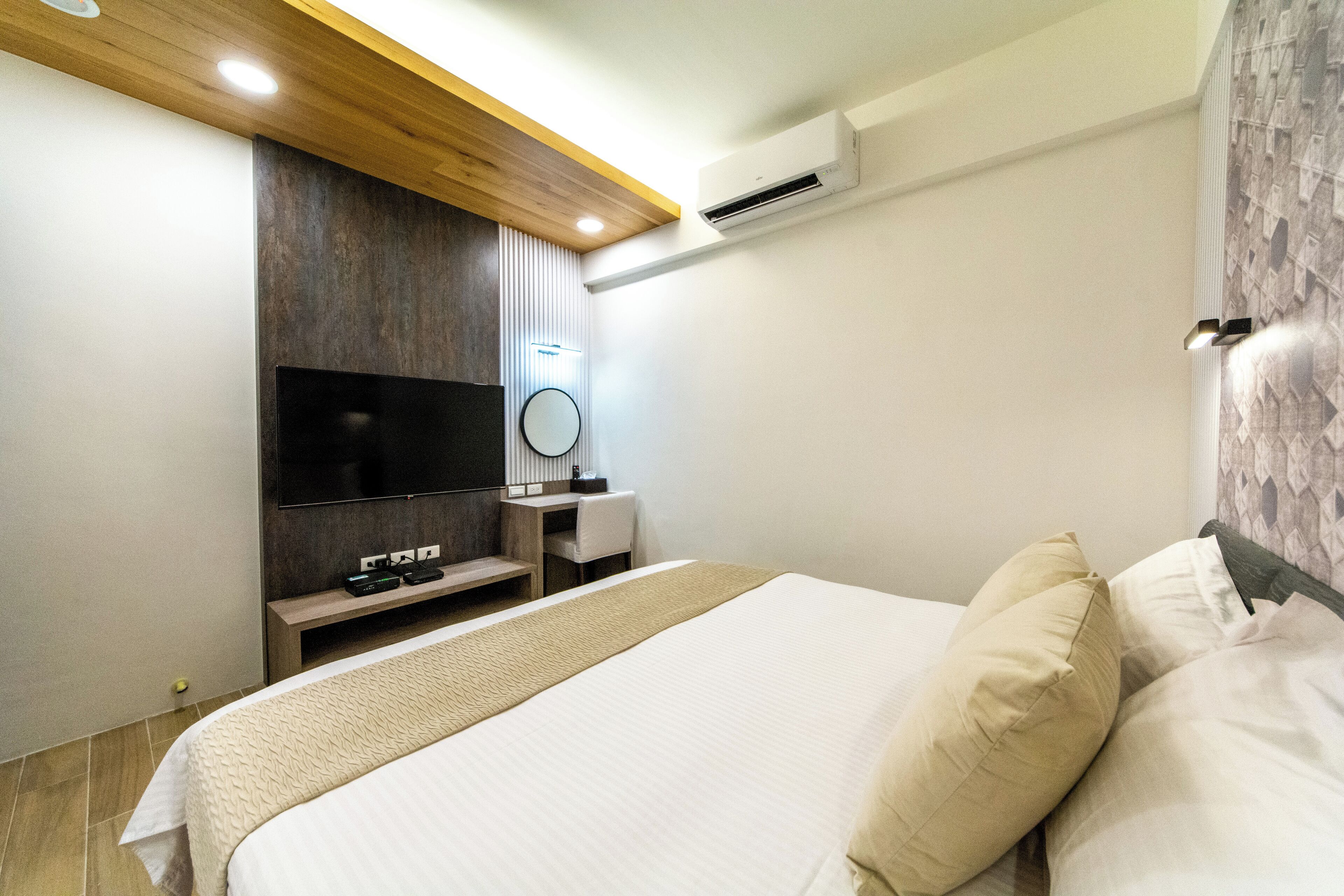 A modern hotel room with a large bed, a flat-screen TV mounted on the wall, a desk with a chair, and an air conditioning unit.