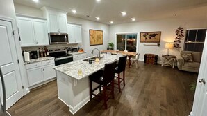 Una cocina moderna con armarios blancos, encimera de granito y una isla central. Hay un comedor con mesa y sillas de madera.