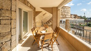 A balcony with a wooden table and chairs, stone walls, and a view of the street.