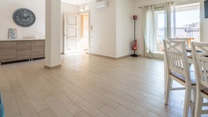 A spacious room with a wooden floor, a dining area with white chairs, a wall clock, and a fire extinguisher.