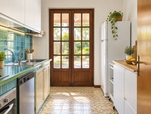 A kitchen with a double door leading to a garden, a sink with utensils, a refrigerator, and a countertop with a plant.