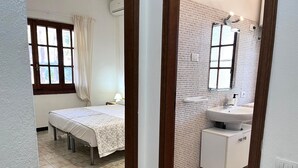 A bedroom with a bed, a nightstand, a window with curtains, and a bathroom with a sink and mirror. - Vacation home Itaca by Interhome (Villasimius)