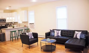 A modern living room with a black leather sofa, two green bar stools, and a small coffee table. A kitchen with white cabinets and a stone backsplash is visible in the background.