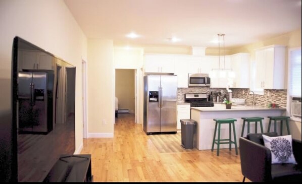 A modern kitchen with stainless steel appliances, a central island with stools, and a hallway leading to another room.