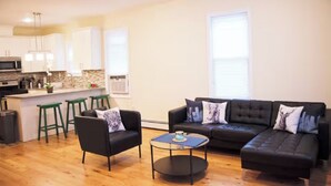A modern living room with a black leather sofa, two green bar stools, and a small coffee table. A kitchen with white cabinets and a stone backsplash is visible in the background. - Harrel Vacation Rentals (Omaha)