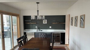 Ein moderner Essbereich mit Holztisch und -stühlen, eine Küche mit dunkler Spritzschutzverkleidung und ein großes Fenster mit Blick ins Freie.