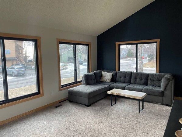Ein Wohnzimmer mit einem Ecksofa, einem Holztisch und großen Fenstern mit Blick auf eine Straße und Gebäude.