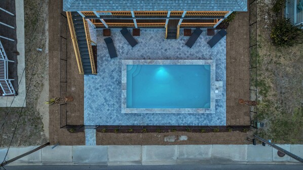 An aerial view of a rectangular pool with a tiled deck, surrounded by a wooden pergola with seating.